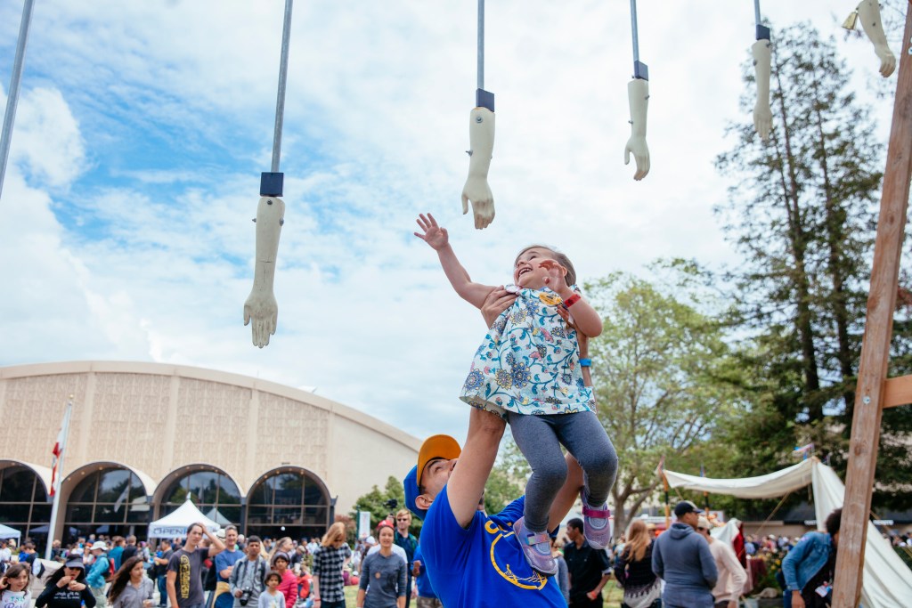 A young girl being held up by her father reaches to grab a dangling hand and give a high five.